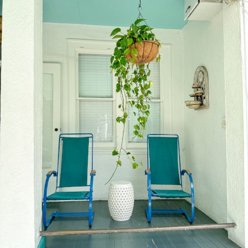 Two blue chairs and a small white table sit on a porch with a hanging plant above. The porch is painted white and turquoise.