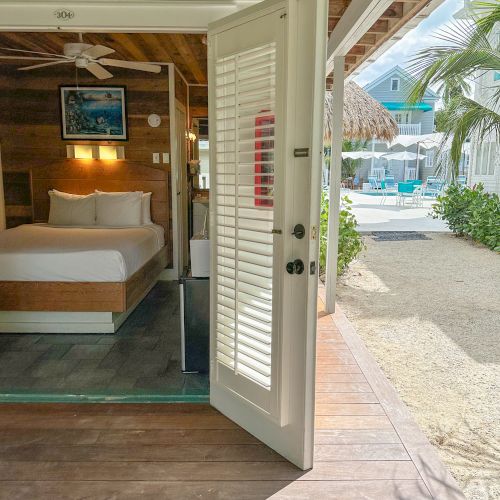 A cozy hotel room with a bed and open door leading to a sunny patio with a pool. Palm trees and blue skies are in the background.