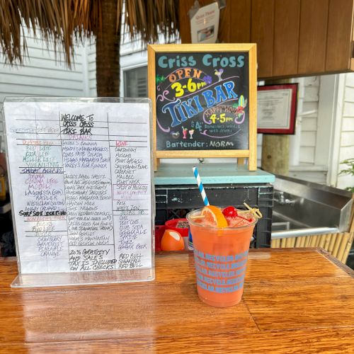 A menu and a colorful drink with a straw and garnishes are on a wooden bar. A sign mentions a tiki bar with specific hours.