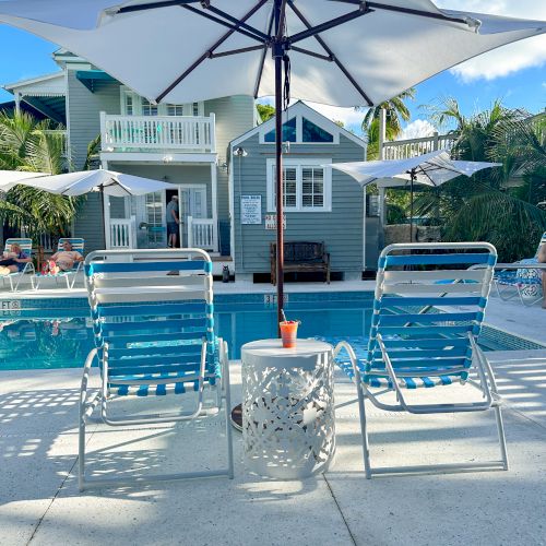 Two lounge chairs under an umbrella beside a pool, with a small table and a house in the background.