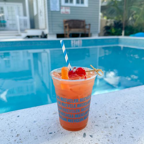 A tropical cocktail with fruit garnishes and a striped straw sits by a poolside. A house and a bench are visible in the background.