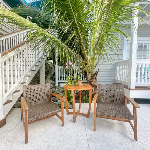 Two wicker chairs and a small table under a large palm plant, next to a staircase and white railing.