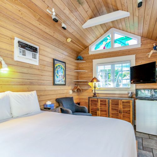 A cozy wooden bedroom with white bedding, wall art, a TV, mini fridge, and seating, well-lit by natural light through windows.