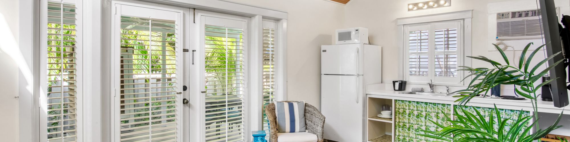 A cozy room with a high wooden ceiling, French doors, a kitchenette, and tropical decor, featuring a ceiling fan and tiled floor.