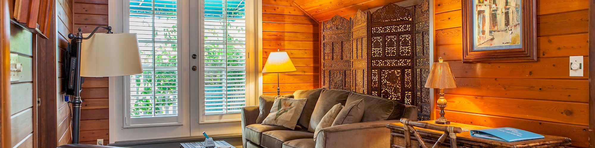 A cozy wooden living room with a sofa, armchair, desk, and lamps. Artwork hangs on the wall, and windows with blinds bring in natural light.