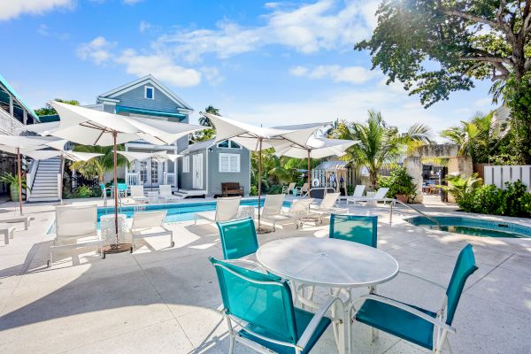 Outdoor pool area with teal chairs, white umbrellas, and a bright house surrounded by trees and plants under a clear blue sky.