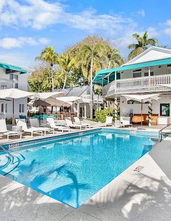 A sunny hotel-style courtyard with a rectangular blue pool, white poolside chairs, sun umbrellas, and pastel two-story buildings with teal awnings.